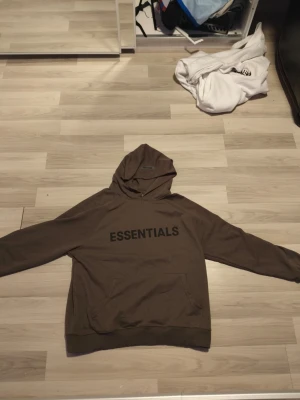 Brun Essentials hoodie med huva - Säljer en brun hoodie från Essentials med stor svart logga på bröstet. Hoodien har en klassisk huva och magficka framtill. Tillverkad i mjukt bomullsmaterial som är perfekt för chill dagar. Enkel och stilren design som passar till det mesta.