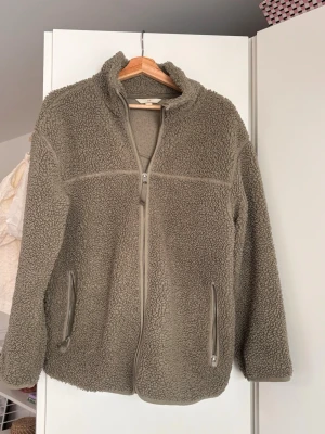 Oliv/pistage-grön teddyjacka med dragkedja - Mysig teddyjacka med hög krage och hel dragkedja framtill. Jackan har två sidofickor med dragkedja och är gjord i fluffigt teddy-material som ger en avslappnad och trendig look. Perfekt för lager-på-lager och chill dagar.