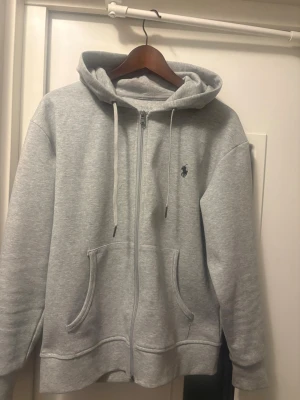Grå zip hoodie från Polo Ralph Lauren - Snygg grå hoodie med dragkedja från Polo Ralph Lauren. Klassisk design med huva och vita dragsnören, helt ny, storlek S. Hör av vid intresse!!☺️
