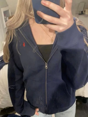 Mörkblå hoodie från Polo Ralph Lauren - Mörkblå hoodie med dragkedja från Polo Ralph Lauren. Klassisk design med huva, fickor framtill och röd broderad logga på bröstet. Tillverkad i mjukt bomullsmaterial som är skönt att bära. Perfekt för en avslappnad och stilren look❤️