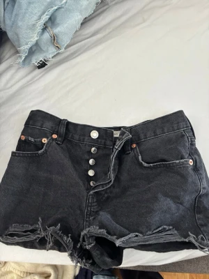 Svarta högmidjade jeansshorts - Säljer ett par svarta högmidjade jeansshorts från Levi's med råa, fransiga kanter och knappgylf. Klassisk femficksmodell i denim som ger en cool och avslappnad vibe. Perfekta till sommaren och lätta att matcha med olika stilar.