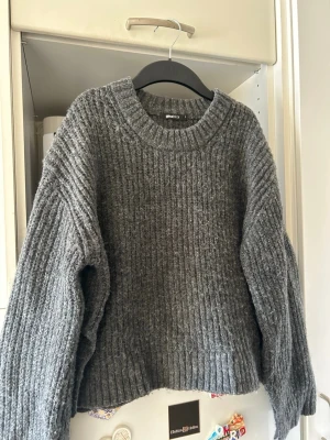 Grå stickad tröja från Gina Tricot - Mysig grå stickad tröja från Gina Tricot med bred ribbstickning och rund hals. Tröjan har en avslappnad passform och långa ärmar, perfekt för kyliga dagar. Materialet känns mjukt och värmande, och den neutrala färgen gör den enkel att matcha.