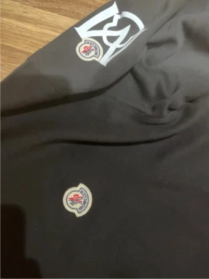 Svart sweatshirt från Moncler - Svart sweatshirt från Moncler med klassisk rund halsringning. Tröjan har Moncler-logga på bröstet och en stor vit logga på ena ärmen. Tillverkad i mjukt material som passar perfekt till en avslappnad stil.