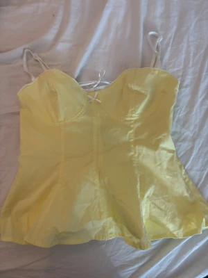 Gul korsett-topp med snörning - Säljer en ljusgul korsett-topp med tunna axelband och liten rosett framtill. Toppen har en figurnära passform och snörning i ryggen som ger en snygg siluett. Perfekt för dig som vill ha en trendig och feminin look.