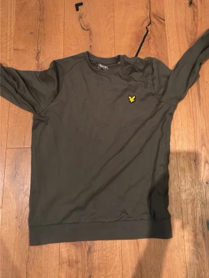 Mörkgrön sweatshirt från Lyle & Scott - Mörkgrön sweatshirt från Lyle & Scott med klassisk rund halsringning och gul logga broderad på bröstet. Tröjan har långa ärmar och ribbade muddar vid ärmslut och nederkant. Perfekt för en avslappnad och stilren look.