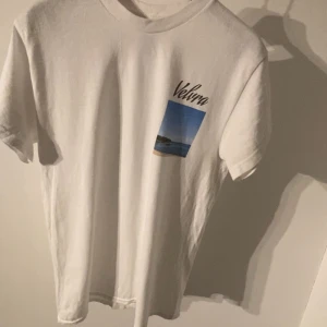 Vit Velvra t-shirt med tryck - Vit t-shirt från Velvra med ett litet tryck på bröstet som föreställer ett landskapsfoto och texten 'Velvra'. Klassisk passform och rund halsringning. Perfekt till jeans eller shorts för en clean och enkel stil.