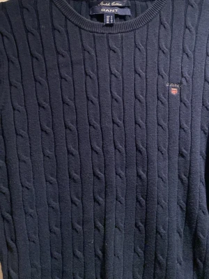 Mörkblå kabelstickad tröja Gant - Klassisk mörkblå kabelstickad tröja från Gant med rund halsringning och diskret logga på bröstet. Tröjan har långa ärmar och ribbade muddar vid ärmslut och nederkant. Perfekt för en stilren och tidlös look. Pris går att diskutera