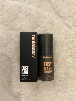 Huda Beauty Easy Bake Setting Spray - Fräsch face mist från Huda Beauty i modellen Easy Bake Setting Spray. Kommer i en svart, kompakt sprayflaska med matchande svart förpackning. Perfekt för att fixera makeup och ge en naturlig finish. Innehåller 30 ml. Oanvänd, helt ny
