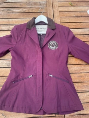 Vinröd kavaj från PS of Sweden - Snygg vinröd kavaj från PS of Sweden med brunt krage och muddar. Kavajen har två dragkedjefickor framtill och en rund logga på bröstet. Figursydd modell. Helt ny inköpt från Falsterbo 2024 så endast provas