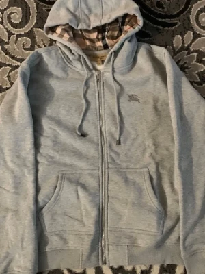 Grå zip hoodie från Burberry - Snygg grå zip hoodie från Burberry med klassiskt rutigt foder i huvan och broderad logga på bröstet. Hoodien har dragsko i huvan, två fickor fram och är tillverkad i mjuk bomullsblandning. Perfekt för en chill och stilren look.