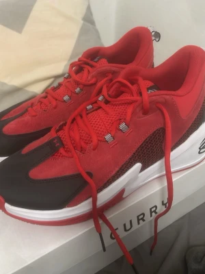 Röda Curry basket sneakers från Under Armour - Säljer ett par snygga röda Curry basket sneakers från Under Armour. Skorna har meshpaneler för bra ventilation, svarta och vita detaljer samt en slitstark yttersula med mönster för extra grepp. Perfekta för basket eller streetwear.