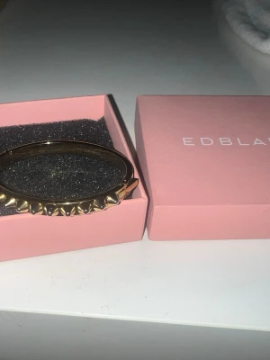 Guldigt armband från Edblad - Snyggt armband i guldfärgad metall från Edblad. Armbandet har en stel form och är dekorerat med coola nitar runt hela. Perfekt accessoar för att ge din outfit en edgy touch. Levereras i original rosa ask.