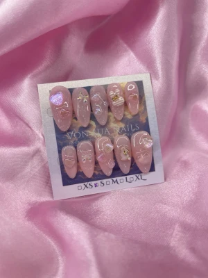 RosiePop (S);Medium Almond - Säljer ett set handgjorda  press-on naglar i en mjuk rosa glittrig nyans. Naglarna har en medium almondform och dekoreras med vita linjer, hjärtan, guldiga detaljer och skimrande effekter. Perfekt för dig som vill ha en söt och trendig look med extra bling.S: 15 mm, 11,5 mm, 12 mm, 11 mm, 9 mm 