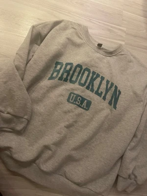 Grå sweatshirt med BROOKLYN-tryck - Säljer en grå sweatshirt med texten BROOKLYN U.S.A. i grönt framtill. Tröjan har rund halsringning, långa ärmar och ribbade muddar vid ärmslut och nederkant. Perfekt för en avslappnad och sportig stil.