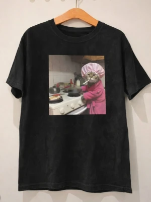 Svart t-shirt med katt i kökstryck - Cool svart t-shirt med ett tryck av en katt i rosa morgonrock och kockmössa som lagar mat. T-shirten har normal passform och korta ärmar. Perfekt för dig som gillar humoristiska motiv och vill sticka ut lite extra.