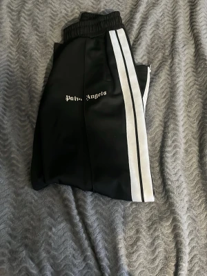 Palm Angels svarta byxor - Säljer ett par svarta mjukisbyxor från Palm Angels med vita dubbla sidoremsor och logga på vänster ben. Byxorna är i använt skick och storlek M. Passar ca 173-180. Pris och frågor hör av er!