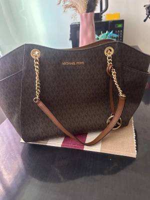 Brun handväska från Michael Kors - Snygg brun handväska från Michael Kors med guldfärgade kedjedetaljer och logotyp framtill. Väskan har ett klassiskt MK-mönster, bruna läderhandtag och rymlig form. Perfekt för dig som gillar stilrena accessoarer med lyxig känsla.