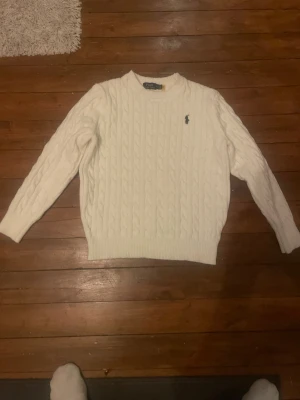 Vit kabelstickad tröja från Polo Ralph Lauren - Säljer en klassisk vit kabelstickad tröja från Polo Ralph Lauren med rund halsringning och den ikoniska lilla loggan broderad på bröstet. Tröjan har långa ärmar och ribbade muddar vid ärmslut och nederkant. Perfekt för en clean och stilren look. Extremt snygg, storlek M men jag skulle säga att den passar ish 170 lång för den är rätt så kort i armarna! Skriv vid frågor! Och pris är diskuterbart😍