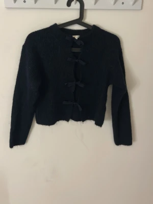 Svart stickad kofta med rosetter - Säljer en mörkblå stickad kofta med långa ärmar och fyra dekorativa rosetter framtill istället för knappar. Kortare modell med rund halsringning. Perfekt för dig som gillar en söt och trendig look.