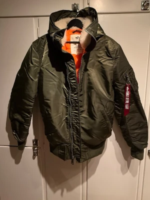 Alpha Industries MA-1 Vinterjacka - Helt nya, aldrig använd bomberjacka från Alpha Industries med huva och orange innerfoder. Jackan har en klassisk utility-ficka på ärmen med röd tag, ribbade muddar och dragkedja framtill. Fodrad med mjuk vit pälsimitation i kragen för extra komfort.