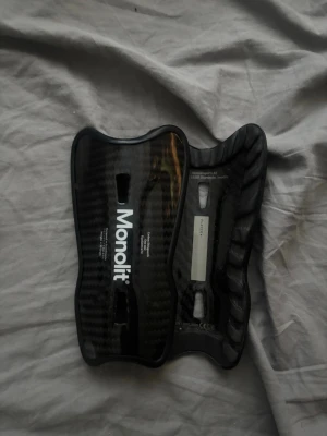 Monolit benskydd i kolfiber 14cm - Säljer ett par svarta benskydd från Monolit, tillverkade i kolfiber med en snygg glansig finish. De har en ergonomisk form för bra passform och är superlätta. Perfekta för dig som vill ha maximalt skydd och stil på planen.