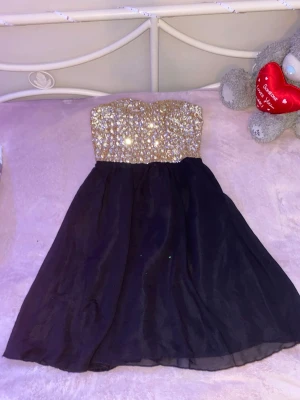 Svart klänning med glittrig överdel Vila - Axelbandslös klänning från Vila med svart, vid kjol och en överdel täckt av guldiga och silvriga paljetter. Perfekt för dig som vill sticka ut med en glittrig look. Klänningen har en smickrande passform och elastisk rygg för extra komfort. FINNS PÅ FLER SIDOR!!