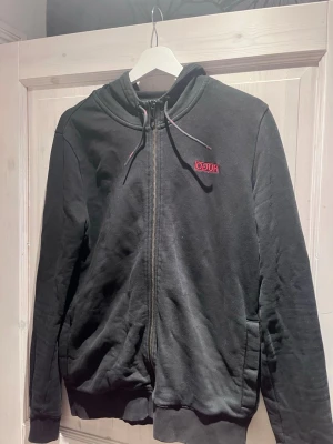 Svart hoodie från Hugo Boss - Snygg svart hoodie från Hugo Boss med dragkedja framtill och liten röd logga på bröstet. Hoodien har huva med dragsko och ribbade muddar vid ärmslut och nederkant. Tillverkad i mjukt bomullsmaterial som känns skönt mot huden.