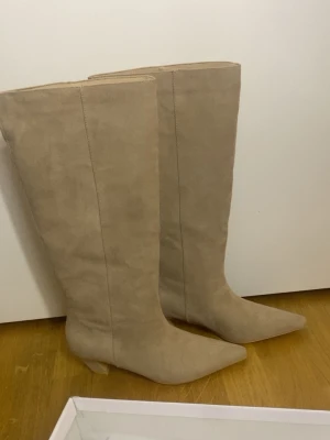 Beige knähöga mockastövlar - Snygga beige stövlar från Nelly i mockaimitation med spetsig tå och låg klack. Endast testade! Nypris 899kr 