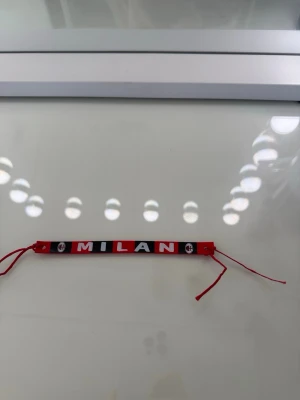 AC Milan armband i rött och svart - Snyggt armband i tyg med AC Milan-tema. Armbandet har röd och svart bakgrund med texten 'MILAN' i vitt samt klubbens logga på sidorna. Justerbara röda band för att passa handleden. Perfekt accessoar för dig som älskar fotboll och vill visa ditt stöd för Milan.