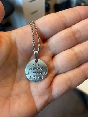 Silverfärgat halsband med textbricka - Runt halsband i silverfärgad metall med en bricka där det står 'She believed she could so she did' ingraverat. Kedjan är av mellanstora länkar och brickan har en enkel, polerad finish. Perfekt för dig som gillar inspirerande smycken.