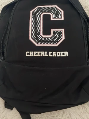 Svart ryggsäck med CHEERLEADER-tryck - Svart cheerleading ryggsäck med rosa diamanter och glitter. Köpt 2024 från Cheer life och har därför tappat lite diamanter. Köpt för 550 men säljer för  320 då diamanter fattas.