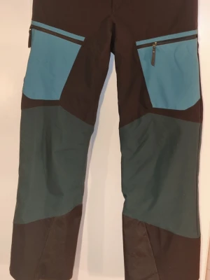 Täckbyxor med blå och gröna detaljer -  Ett par skidbyxor från PeakPerformance i en flerfärgad design. Byxorna har en kombination av svart, turkos och mörkgröna