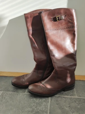 Bruna knähöga stövlar från Esprit - Snygga bruna knähöga stövlar från Esprit i äkta läder. Klassisk design med dekorativt spänne upptill och broguemönster längs sömmarna. Dragkedja på insidan för enkel påtagning och låg klack för bekväm vardagsstil.