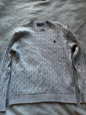 Grå kabelstickad tröja Polo Ralph Lauren - Säljer en grå kabelstickad tröja från Polo Ralph Lauren med klassisk rund hals och diskret broderad logga på bröstet. Tröjan har ribbade muddar vid ärmslut och nederkant, perfekt för dig som gillar stilrena och tidlösa plagg. Skriv vid minsta lilla fundering