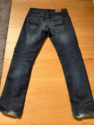  Nudie Jeans - Säljer ett par mörkblå jeans från Nudie Jeans med klassisk femficksdesign och kontrastsömmar. Jeansen har raka ben och är tillverkade i slitstark denim. Perfekt för dig som gillar en tidlös och cool look. Mått : Midja 42 Längd 110 ben öppning 20