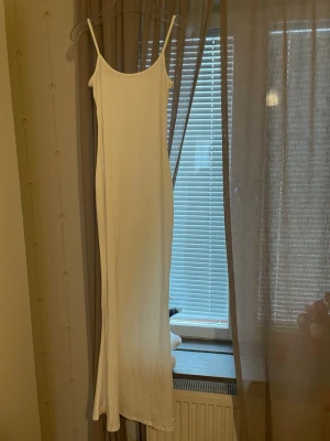 Vit långklänning med tunna axelband - Säljer en enkel och stilren vit långklänning med tunna axelband och rak passform. Klänningen har en minimalistisk look och är perfekt för varma dagar eller som basplagg att styla upp. Materialet känns mjukt och följsamt.