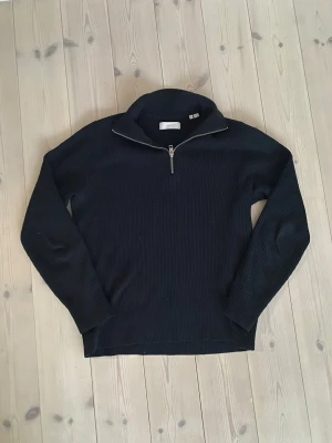 Svart stickad halvzip-tröja - Säljer en svart stickad tröja med halv dragkedja och hög krage. Tröjan har ribbad struktur och långa ärmar, perfekt för lager på lager. Enkel och stilren design som passar till det mesta.