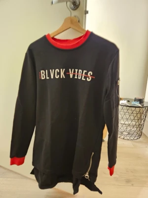 Svart sweatshirt - Cool svart sweatshirt från BLVCK VIBES med tryck framtill och röda detaljer vid hals och ärmslut. Tröjan har dragkedja i sidan. Använd 3-4 gånger, som ny.