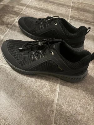 Svarta sneakers från ECCO - Snygga svarta sneakers från ECCO med mesh- och syntetmaterial. Skorna har elastiska snören med snabblås och en sportig, låg profil. Perfekta för dig som gillar stilrena och funktionella skor med bra andningsförmåga. Som nya!