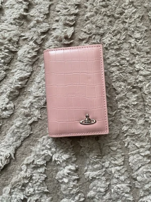 Rosa plånbok från Vivienne Westwood - Snygg rosa plånbok i glansigt material med krokodilpräglat mönster från Vivienne Westwood. Plånboken har det ikoniska silverfärgade Orb-emblemet på framsidan och en stilren, vikbar design. Perfekt för dig som gillar trendiga accessoarer.