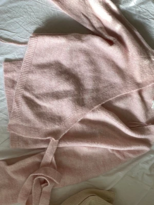 Ljusrosa omlottkofta Gina Tricot - Mysig ljusrosa stickad omlottkofta från Gina Tricot i ullblandning. Koftan har långa ärmar, ribbade muddar och knytband i midjan för en snygg passform. Perfekt för lager på lager och ger en soft vibe till din outfit.