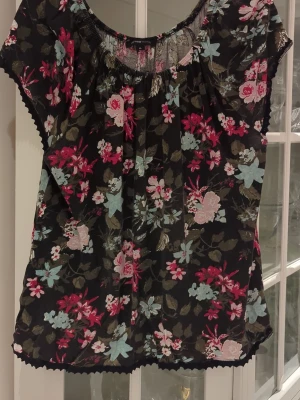 Blommig topp från mywear woman - Söt blommig topp från mywear woman i svart med färgglada blommor i rosa, blått och vitt. Toppen har korta ärmar, rund halsringning med resår och är gjord i ett lätt och luftigt material. Använd