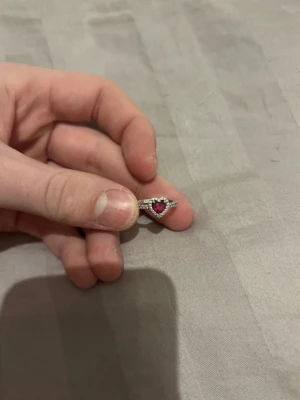 Silverring med hjärtformad röd sten - Snygg silverfärgad ring med en hjärtformad röd sten i mitten, omgiven av små klara stenar som ger extra bling. Ringen har en tunn banddesign med små stenar längs hela bandet för en elegant look. Perfekt för dig som gillar romantiska detaljer och vill sticka ut. Från Pandora