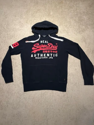 Mörkblå hoodie från Superdry - Snygg mörkblå hoodie från Superdry med stor logga och japansk text i rött och vitt på bröstet. Huva med vita dragsnören och magficka framtill. Röd detalj med siffran 28 på ena ärmen. Mjuk insida i grått tyg, perfekt för chill dagar.