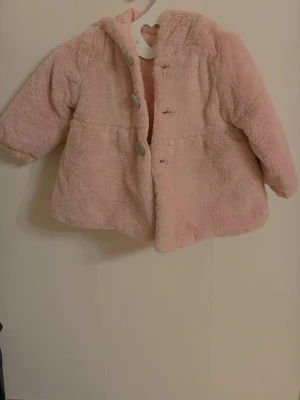 Ljusrosa fuskpälsjacka med huva - Barn  Supersöt ljusrosa pälsjacka i fuskpäls med mjukt quiltat innerfoder. Jackan har huva och stängs med knappar framtill. Perfekt för kalla vinterdagar när du vill vara både varm och stylish.