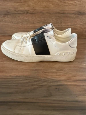Valentino open - Snygga vita sneakers från Valentino med svart detalj på sidan och coola nitar längs hälen. Skorna har klassisk låg profil, vita snören och är tillverkade i läder. Perfekta för dig som vill ha en stilren men edgy look.