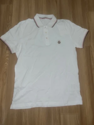 Vit Moncler pikétröja med röd detalj - Klassisk vit pikétröja från Moncler med röd och marinblå rand på krage och ärmslut. Tröjan har knappslå med tre knappar och Moncler-logga broderad på bröstet. Perfekt till en clean och sportig stil.