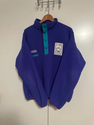 Lila fleece från Columbia Lillehammer 94 - Retro lila fleece från Columbia med tryckknappar i turkos och detaljer från OS Lillehammer 94. Samma tröja som OS-atleterna fick 1994, väldigt unik och få producerade.