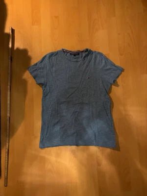 Blå randig t-shirt Tommy Hilfiger - Snygg blå t-shirt från Tommy Hilfiger med tunna vita ränder och klassisk logga på bröstet. Står storlek M men passar bättre som en S. T-shirten har rund halsringning och korta ärmar. Tillverkad i mjuk bomull som känns skön mot huden. Perfekt för en avslappnad och stilren look.