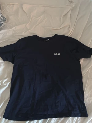 Mörkblå BOSS t-shirt i bomull - Snygg mörkblå t-shirt från BOSS med klassisk passform och vit logga på bröstet. Tillverkad i mjuk bomull och har rund hals samt korta ärmar. Perfekt för en clean och stilren look. Lite mindre i storleken